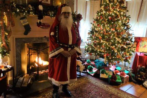 A festive room with a decorated Christmas tree, presents, and a man in a Santa suit standing by a glowing fireplace.
