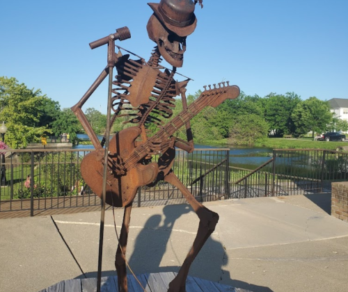 A metal skeleton statue playing guitar on a stage, with a park and pond in the background under a clear blue sky.