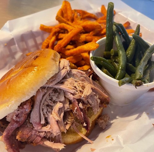 A pulled pork sandwich on a bun with sweet potato fries and green beans on the side, served in a takeout container.