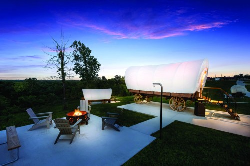 A scenic view of a campsite with a covered wagon, fire pit, and chairs under a colorful sunset sky.