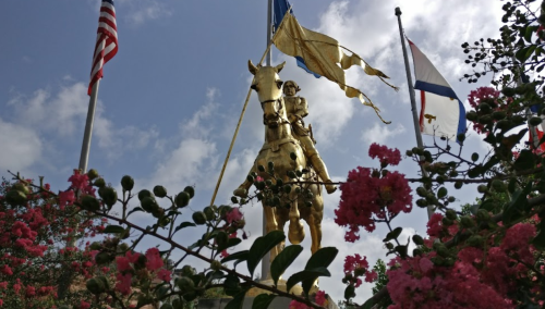 A golden statue of a mounted figure surrounded by flags and blooming pink flowers against a cloudy sky.