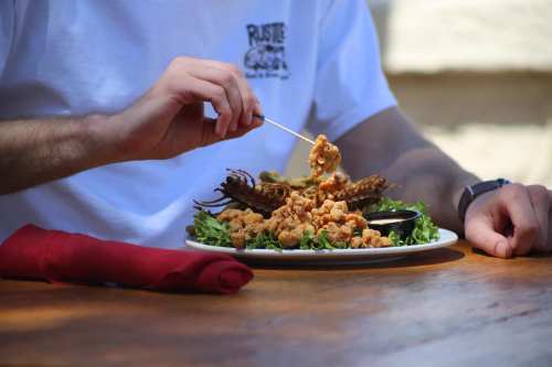 A person holds a skewer while enjoying a plate of fried seafood on a wooden table, with a red napkin nearby.