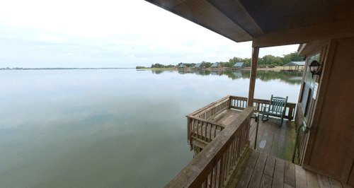 A serene lakeside view from a wooden deck, featuring calm waters and distant cabins under a cloudy sky.