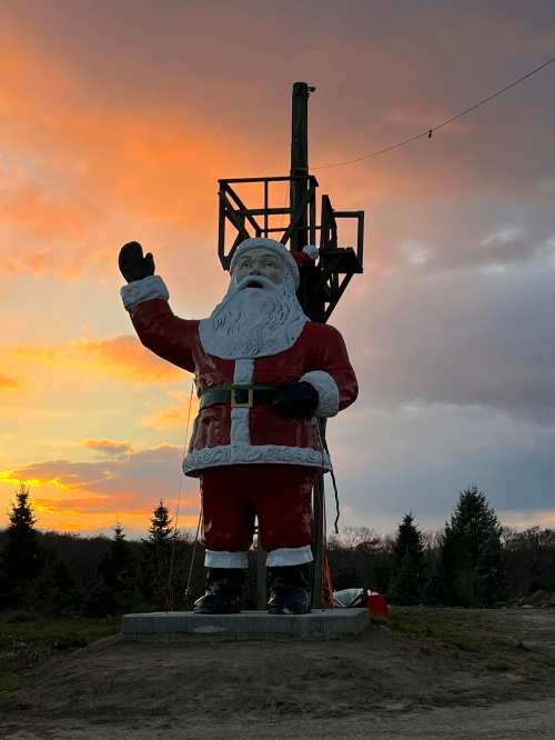 A large Santa Claus statue waves against a colorful sunset, with trees in the background.
