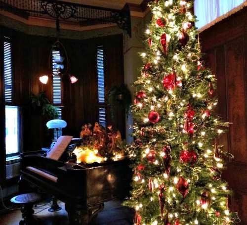 A beautifully decorated Christmas tree with red ornaments stands next to a grand piano in a cozy, festive room.