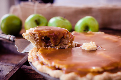 A slice of caramel apple pie on a serving spatula, with green apples in the background.