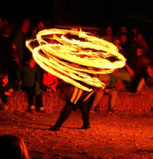 A performer spins flaming poi in front of an audience at a nighttime event, surrounded by hay bales.