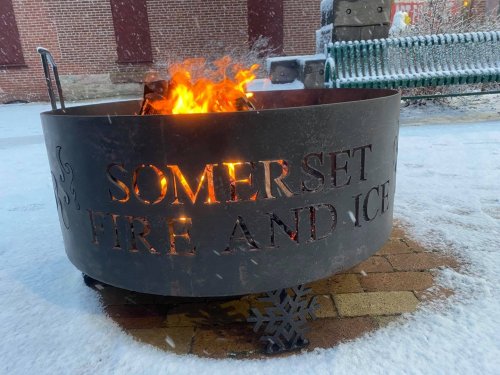 A fire pit with the words "SOMERSET FIRE AND ICE" burning brightly, surrounded by snow and a brick background.