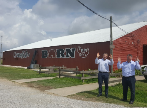 Two men in dress shirts stand outside a red barn with "Crawfish BARN" painted on the side, raising their hands.