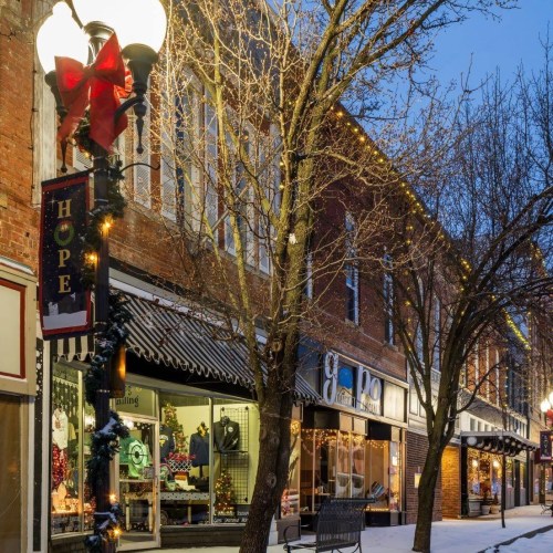 A charming street decorated for the holidays, featuring snow, trees, and festive lights on storefronts.