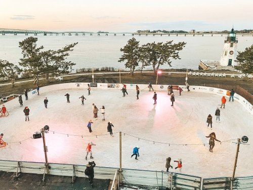 Aerial view of an outdoor ice skating rink with people skating, surrounded by trees and a lighthouse by the water.