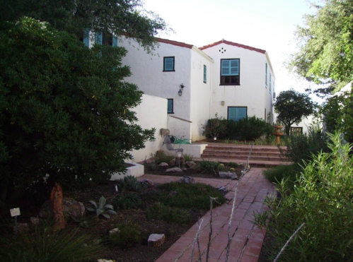 A pathway leads to a white building with blue windows, surrounded by greenery and stone landscaping.