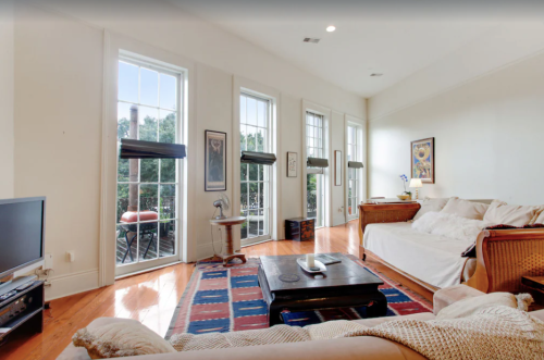 Bright living room with large windows, a cozy sofa, coffee table, and a TV, featuring wooden floors and colorful rugs.
