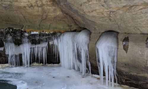 Icicles hanging from a rocky overhang, with a frozen surface below and snow scattered around.