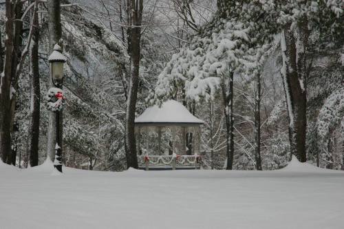 A snow-covered gazebo surrounded by trees, with a lamp post adorned with a festive wreath in a winter landscape.