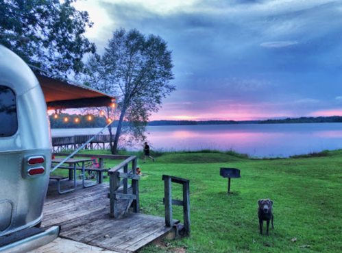 A silver Airstream trailer by a lake at sunset, with a dog and picnic area in the foreground.
