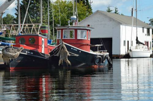 Two fishing boats with red and black colors docked by a white building, reflecting in calm water under a clear sky.