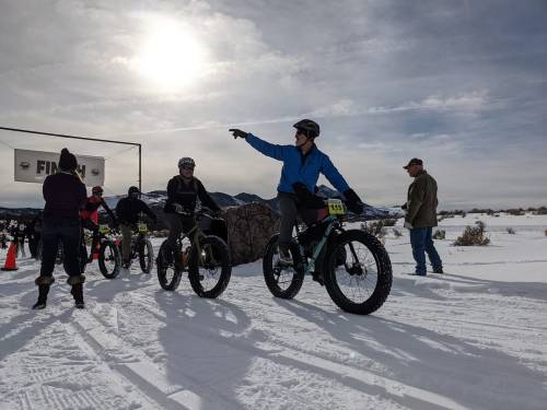 A group of cyclists in winter gear prepares for a race on snow, with one pointing towards the starting line.