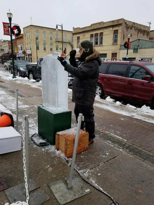 A person in winter clothing carves an ice block on a snowy street, with festive decorations in the background.