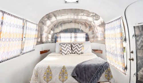 Cozy interior of a vintage Airstream trailer with a bed, decorative pillows, and patterned curtains.