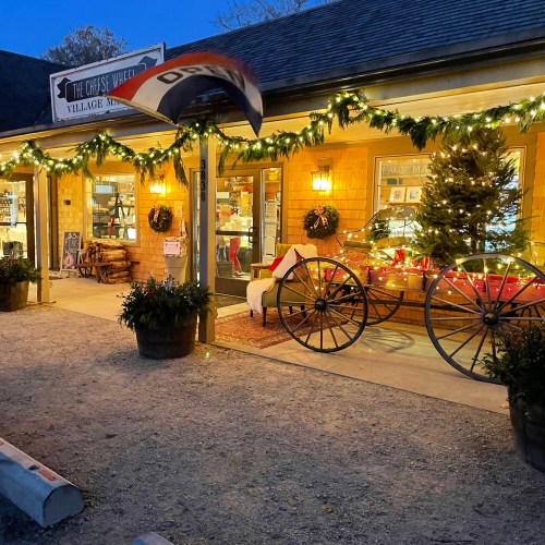 A charming shop exterior decorated with lights and holiday decor, featuring a wagon wheel and potted plants.