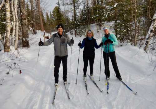 Three people cross-country skiing on a snowy trail surrounded by trees.