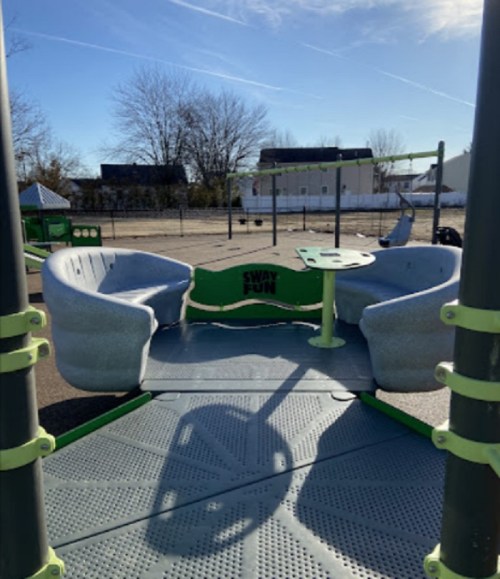 A playground with two curved seats and a central table, surrounded by a fenced area and trees in the background.