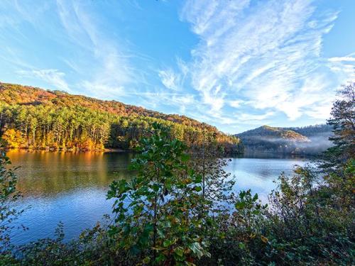 A serene lake surrounded by autumn-colored trees and rolling hills under a blue sky with wispy clouds.