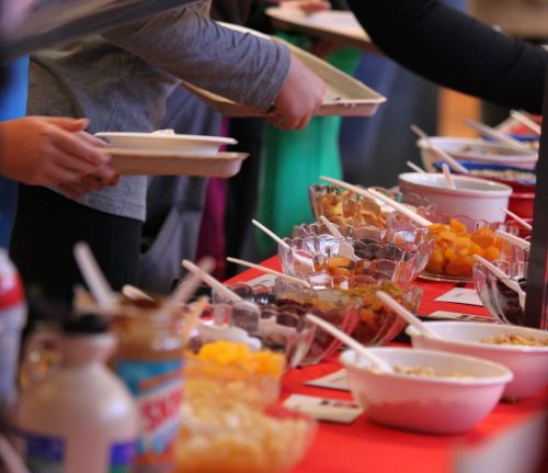 A buffet table with various bowls of food, as people serve themselves with trays and utensils.