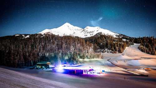 A snowy mountain landscape at night, illuminated by colorful lights, with a starry sky above.