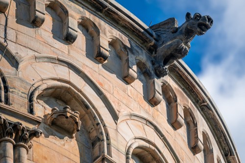 Close-up of a stone building featuring intricate arches and a gargoyle perched on the edge against a blue sky.