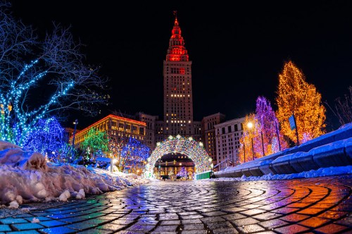 A vibrant cityscape at night, featuring a tall building lit in red, surrounded by colorful holiday lights and snow.