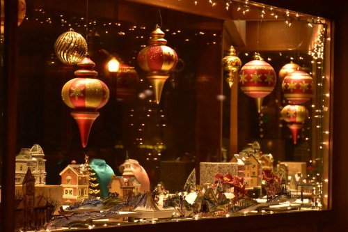 A festive window display featuring colorful ornaments and miniature holiday village decorations.