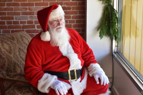 A jolly Santa Claus in a red suit and hat sits on a chair by a window, smiling warmly.