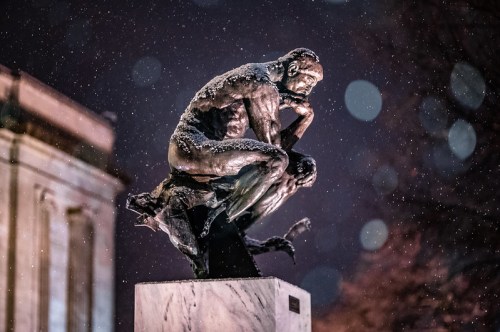 A bronze sculpture of a contemplative figure, partially covered in snow, illuminated at night with a blurred background.
