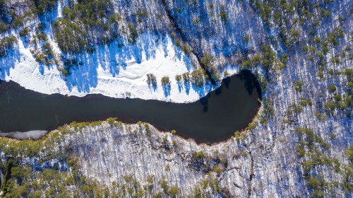Aerial view of a winding river surrounded by snow-covered land and trees in a winter landscape.