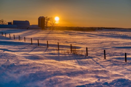 A serene winter landscape at sunset, featuring a snowy field, a barn, and a silhouetted tree against a colorful sky.