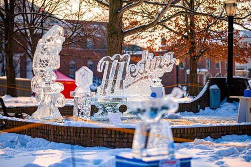 Ice sculptures in a snowy park, featuring the word "Medina" and other artistic designs, with trees in the background.