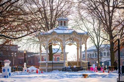 A snow-covered gazebo in a park, surrounded by trees and festive decorations, with a winter landscape in the background.