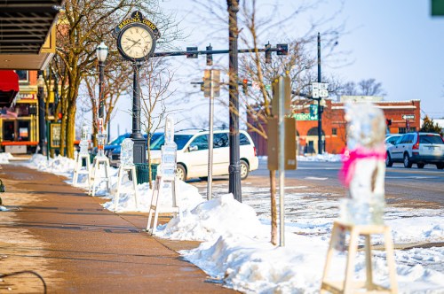 A snowy street scene with a clock, parked cars, and ice sculptures lining the sidewalk.