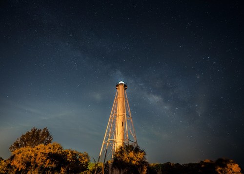 A tall lighthouse illuminated under a starry night sky, with the Milky Way visible in the background.