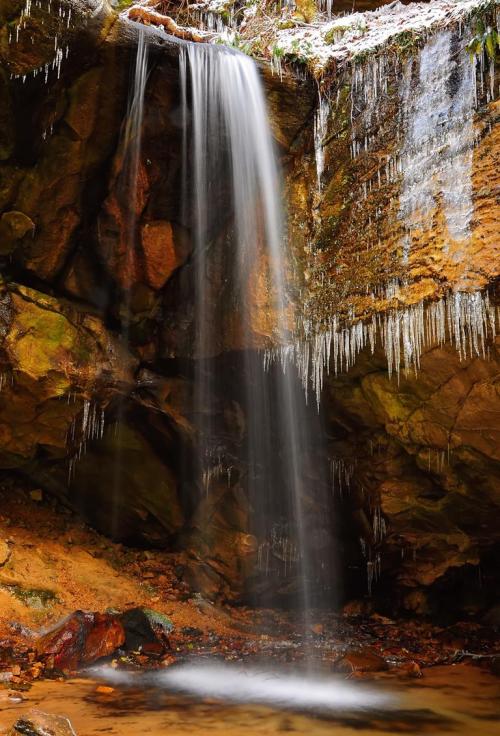 A serene waterfall cascades over rocks, surrounded by icicles and a tranquil pool below.