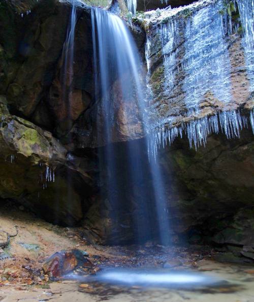 A serene waterfall cascades over rocky cliffs, surrounded by icicles and a tranquil pool below.