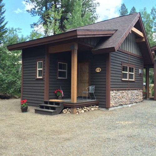 A small, dark wooden cabin with a stone foundation, surrounded by trees and gravel, featuring a porch and steps.