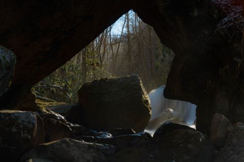 A serene view of a waterfall framed by large rocks and trees, with soft sunlight filtering through.