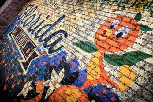 Colorful mural featuring an orange character and the text "Florida" on a brick wall, celebrating local produce.