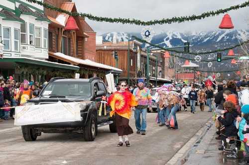 A festive parade with colorful costumes, a decorated truck, and spectators along a street with mountains in the background.