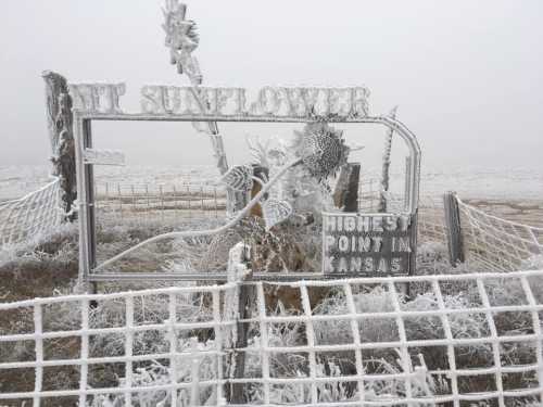 A frosty sign reading "Highest Point in Kansas" with a sunflower design, surrounded by a foggy, icy landscape.