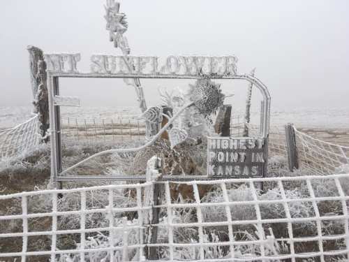 A frosty sign reading "Highest Point in Kansas" with a sunflower design, surrounded by a foggy, icy landscape.