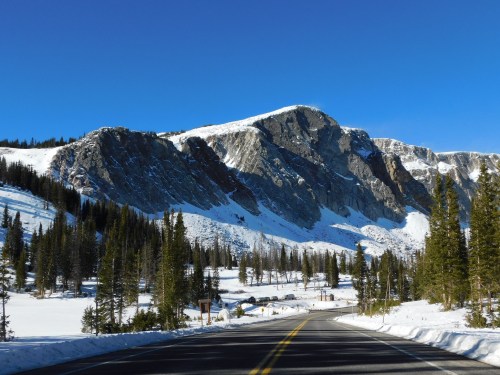 A snow-covered mountain rises above a winding road, surrounded by evergreen trees under a clear blue sky.